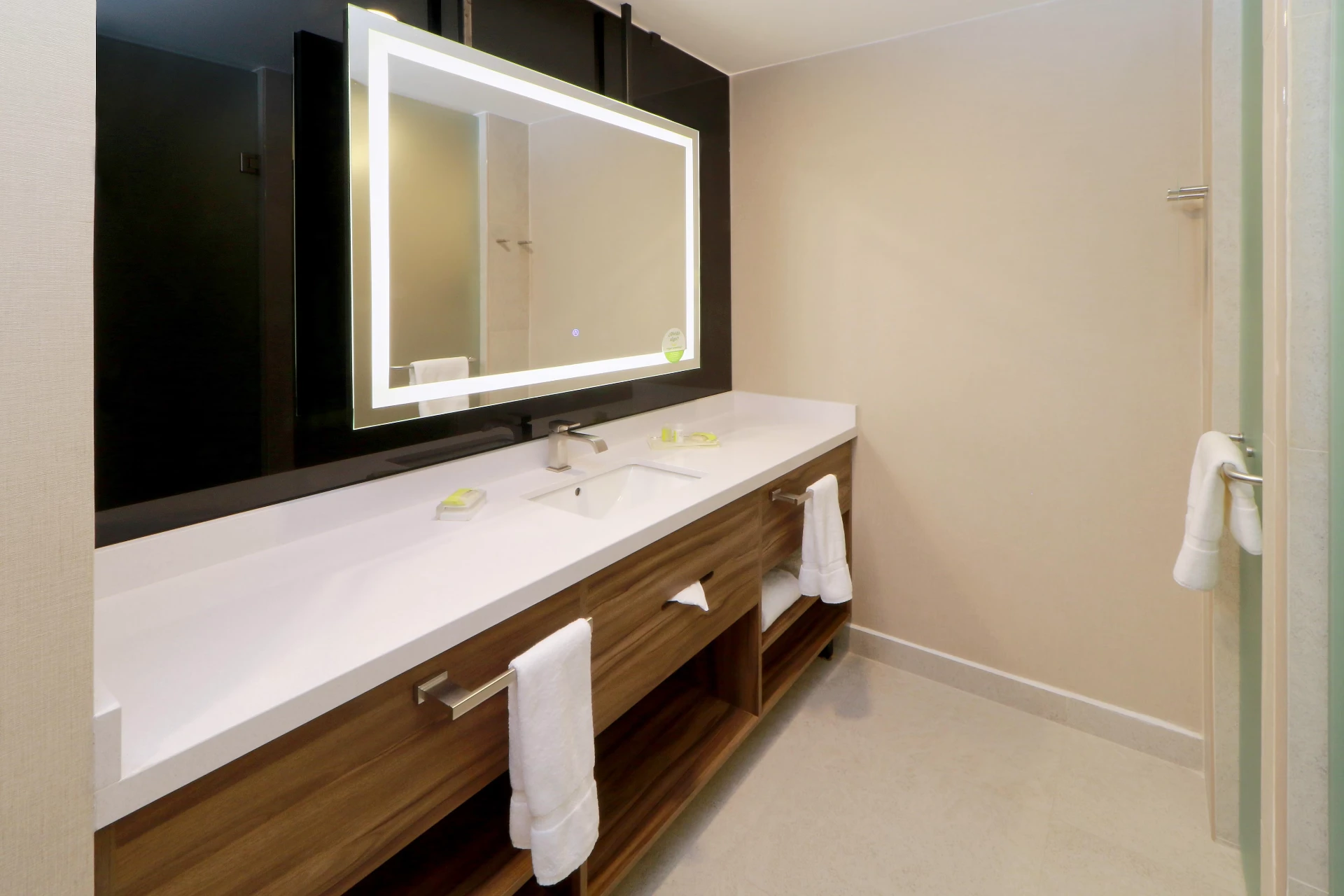 Baño de hotel con espejo LED iluminado sobre lavabo de superficie sólida y mueble de madera