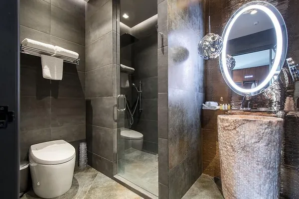Baño de hotel boutique con espejo LED ovalado iluminado sobre lavabo de piedra y ducha de cristal